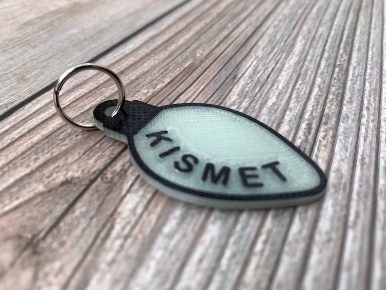 Op de afbeelding: Een witte lampvormige sleutelhanger met een zwarte omtrek en het woord "KISMET" in het zwart gedrukt.