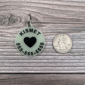 May include: A round, glow-in-the-dark pet ID tag with the name "Kismet" and phone number "555-555-5555" printed in black. A black heart is in the centre of the tag.