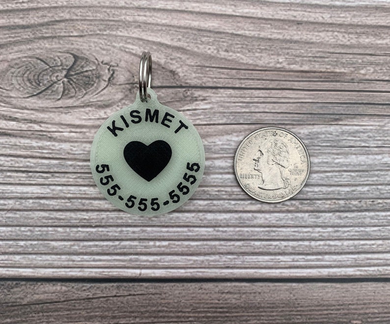 May include: A round, glow-in-the-dark pet ID tag with the name "Kismet" and phone number "555-555-5555" printed in black. A black heart is in the centre of the tag.