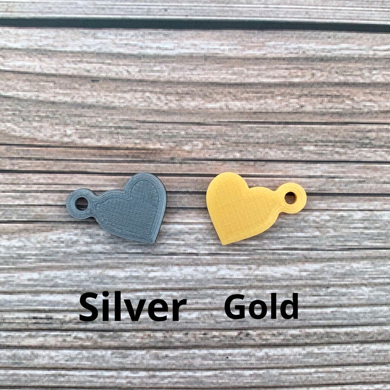 May include: Two heart-shaped charms, one silver and one gold, with a small loop for attaching to a necklace or bracelet. The charms are on a wooden background with the words "Silver" and "Gold" written below them.