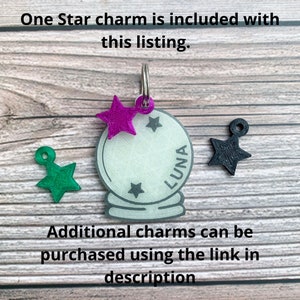 May include: A white glass charm with the word "LUNA" inscribed on it. The charm is attached to a silver ring and has a purple star charm attached to it. There are two other star charms, one green and one black, on a wooden surface.