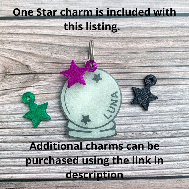 May include: A white glass charm with the word "LUNA" inscribed on it. The charm is attached to a silver ring and has a purple star charm attached to it. There are two other star charms, one green and one black, on a wooden surface.