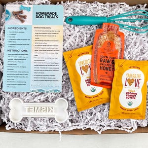 May include: A gift box containing ingredients and instructions for homemade dog treats. Includes peanut butter packets, honey, a whisk, and a bone-shaped cookie cutter. The box is filled with white paper shreds.