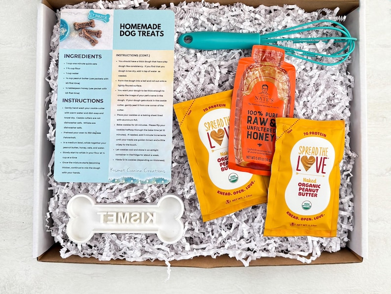 May include: A gift box containing ingredients and instructions for homemade dog treats. Includes peanut butter packets, honey, a whisk, and a bone-shaped cookie cutter. The box is filled with white paper shreds.