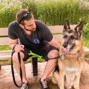 May include: A man wearing a black t-shirt and sunglasses sits on a bench with a German Shepherd dog. The dog is wearing a green leash with the name "Rufus" printed on it.