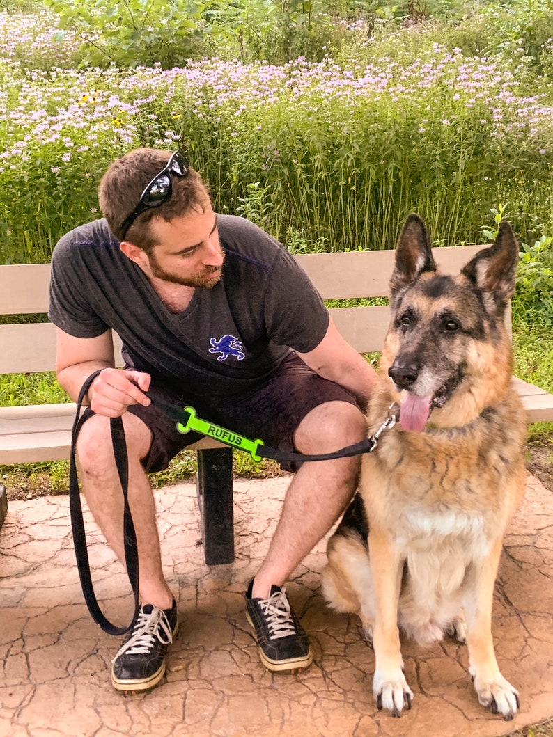 May include: A man wearing a black t-shirt and sunglasses sits on a bench with a German Shepherd dog. The dog is wearing a green leash with the name "Rufus" printed on it.