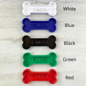 May include: A collection of colorful dog bone-shaped pet tags. The tags are in white, blue, black, green, and red. Each tag has the name "KISMET" engraved on it. The tags are arranged vertically against a light gray background.