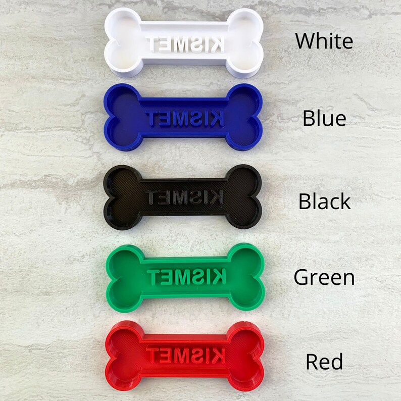 May include: A collection of colorful dog bone-shaped pet tags. The tags are in white, blue, black, green, and red. Each tag has the name "KISMET" engraved on it. The tags are arranged vertically against a light gray background.