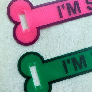 May include: Two dog bone-shaped tags, one pink and one green, with the text "I'M SO" printed on them. The tags have a hole in the middle for attaching to a collar.