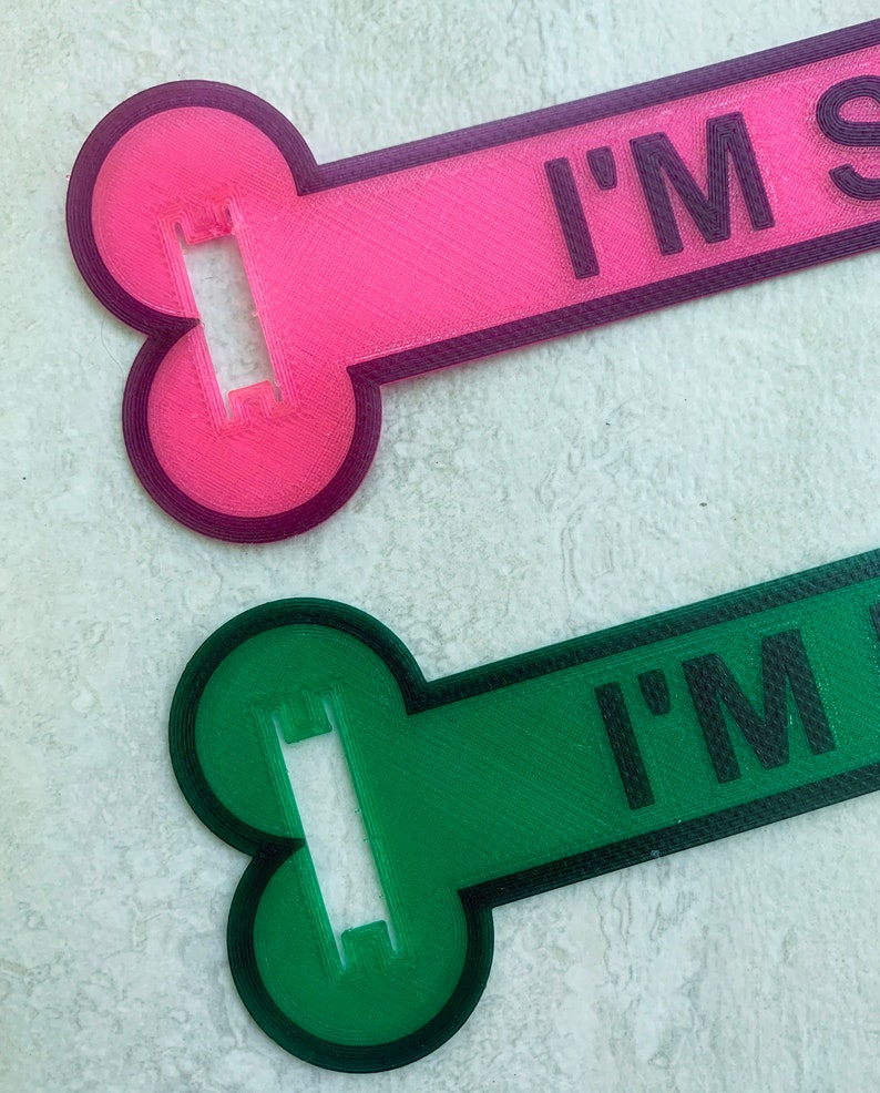 May include: Two dog bone-shaped tags, one pink and one green, with the text "I'M SO" printed on them. The tags have a hole in the middle for attaching to a collar.