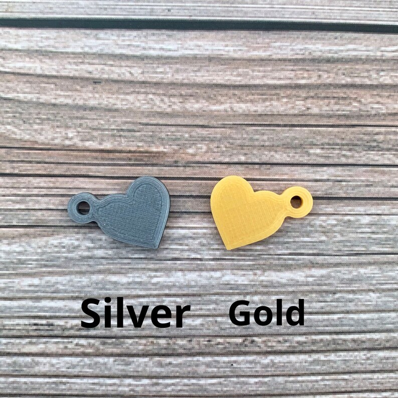May include: Two heart-shaped charms, one silver and one gold, with a small loop for attaching to a necklace or bracelet. The charms are on a wooden background with the words "Silver" and "Gold" written below them.