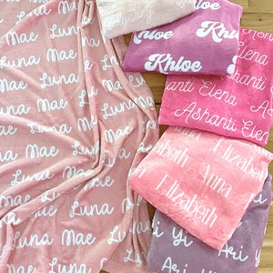 May include: A collection of personalized blankets in various shades of pink and purple. Each blanket features a name in a white, cursive font. The blankets are soft and plush, perfect for a baby or child.