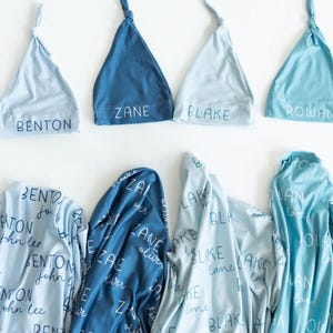 May include: A selection of baby blankets and matching knotted hats in shades of blue. The hats are embroidered with names such as "Arthur," "Benton," and "Zane." The blankets display the same names in a repeating pattern.
