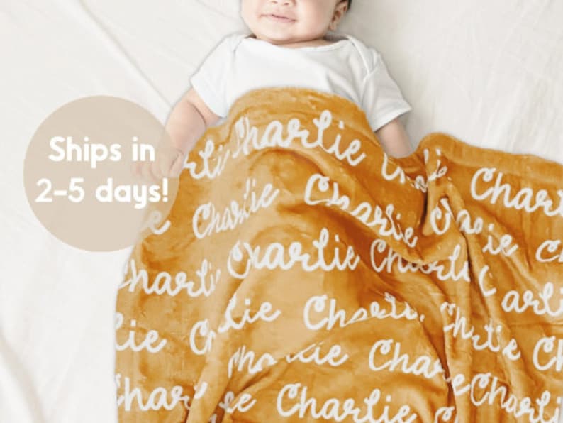 May include: Mustard-colored baby blanket with the name "Charlie" in white script repeated across the surface. The blanket is soft and plush, suitable for a baby. The blanket is draped over a baby wearing a white onesie.