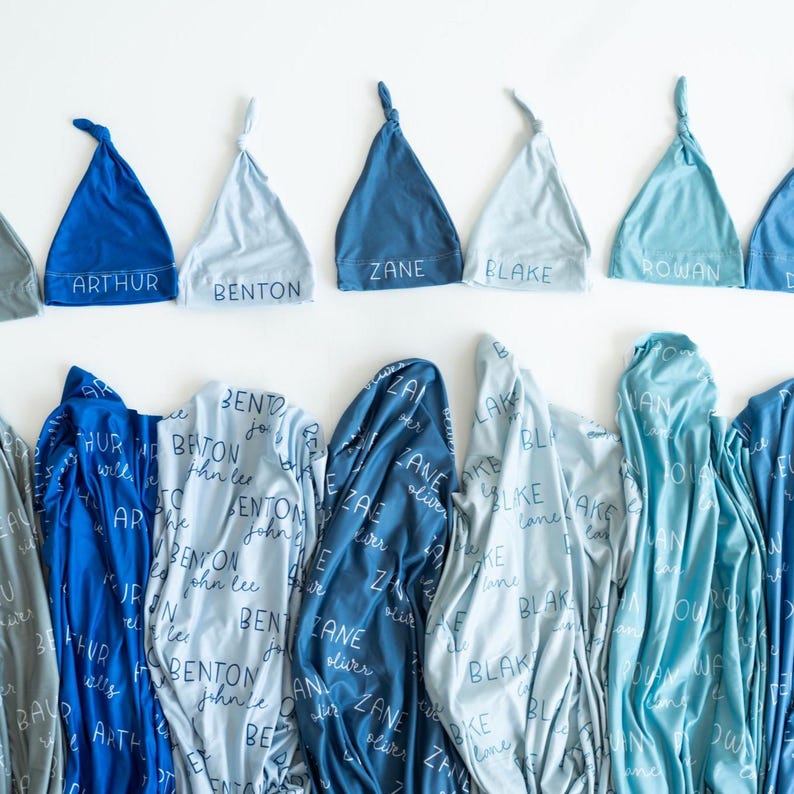 May include: A selection of baby blankets and matching knotted hats in shades of blue. The hats are embroidered with names such as "Arthur," "Benton," and "Zane." The blankets display the same names in a repeating pattern.