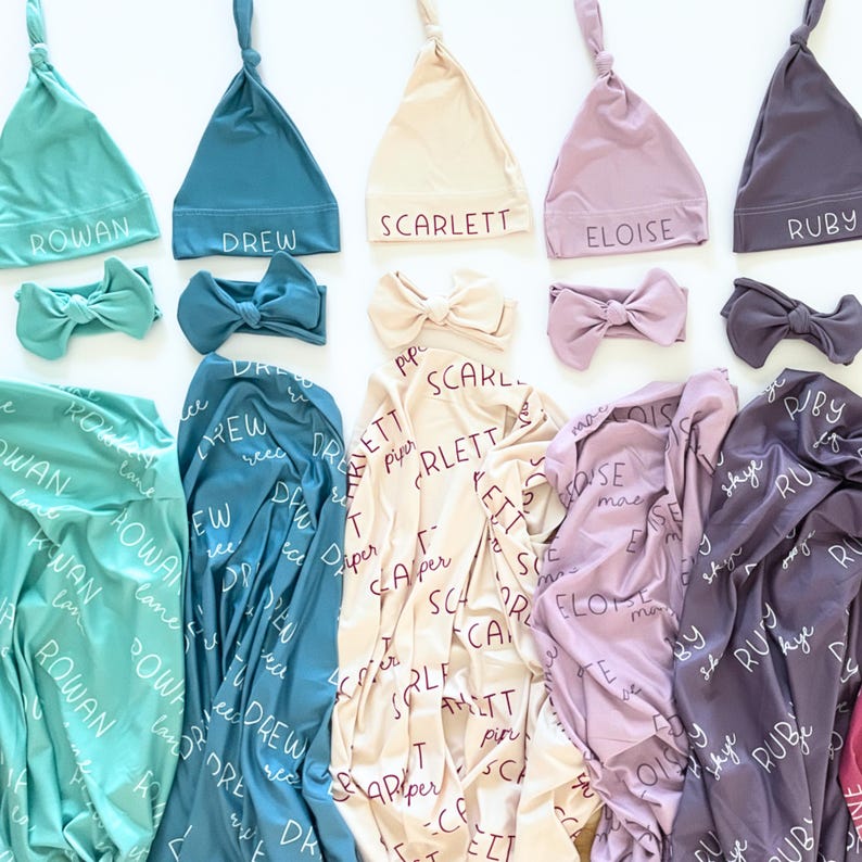 May include: A selection of baby accessories in shades of teal, blue, cream, lavender, and purple. The set includes knotted hats, headbands, and swaddling blankets. Each item is personalised with a name in a script font.