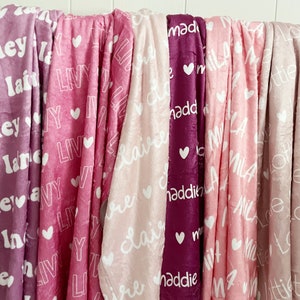 May include: A collection of soft, personalized blankets in various shades of pink and purple. Each blanket features a name in white script and a heart design. The blankets are made of a plush material.