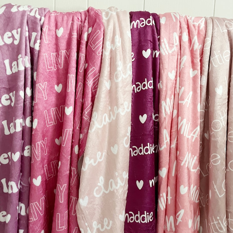 May include: A collection of soft, personalized blankets in various shades of pink and purple. Each blanket features a name in white script and a heart design. The blankets are made of a plush material.