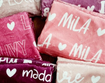 May include: A stack of pink, purple, and burgundy fleece blankets with white embroidered hearts and names, including "Lottie", "Mila", "Ivy", and "Maddie".