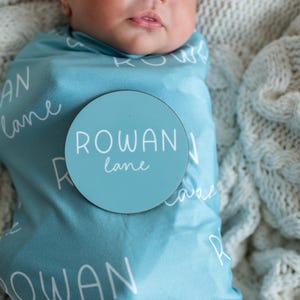 May include: A baby swaddled in a light blue blanket, patterned with the name "Rowan Lane". A circular sign with the name "Rowan Lane" is placed on the blanket. The baby rests on a white knitted blanket.