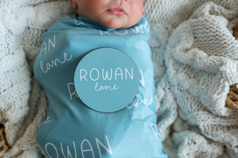 May include: A baby swaddled in a light blue blanket, patterned with the name "Rowan Lane". A circular sign with the name "Rowan Lane" is placed on the blanket. The baby rests on a white knitted blanket.
