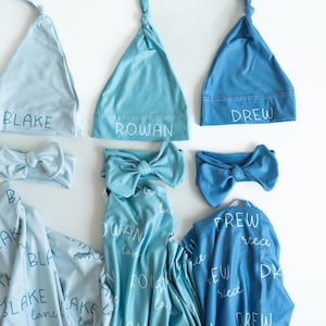 May include: A selection of baby clothing in shades of blue, including knotted hats, headbands with bows, and swaddling blankets. The names "BLAKE", "ROWAN", and "DREW" are printed on the hats. The blankets have the names printed on them as well.