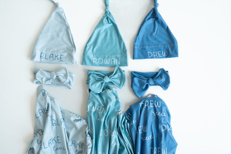 May include: A selection of baby clothing in shades of blue, including knotted hats, headbands with bows, and swaddling blankets. The names "BLAKE", "ROWAN", and "DREW" are printed on the hats. The blankets have the names printed on them as well.