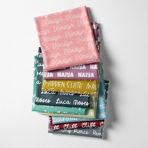 Peut inclure: Une pile de carr&eacute;s de tissu de diff&eacute;rentes couleurs, chacun imprim&eacute; avec un nom diff&eacute;rent en &eacute;criture blanche. Les couleurs incluent le rose, le sarcelle, le violet et l'or. Les noms sont &eacute;crits en motif r&eacute;p&eacute;t&eacute; sur le tissu.