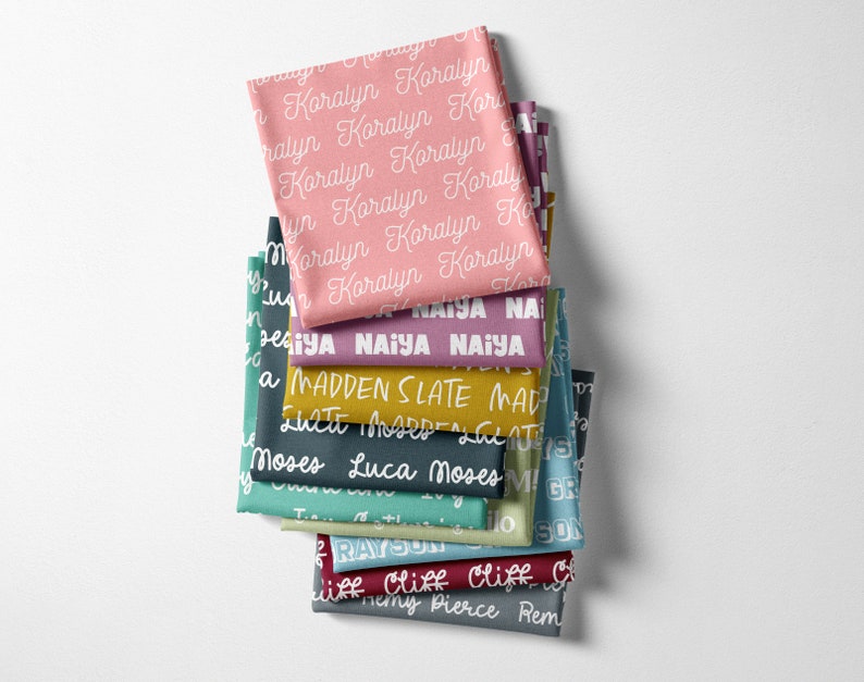 Peut inclure: Une pile de carr&eacute;s de tissu de diff&eacute;rentes couleurs, chacun imprim&eacute; avec un nom diff&eacute;rent en &eacute;criture blanche. Les couleurs incluent le rose, le sarcelle, le violet et l'or. Les noms sont &eacute;crits en motif r&eacute;p&eacute;t&eacute; sur le tissu.
