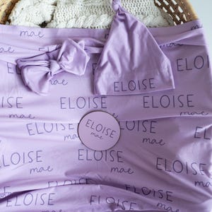 May include: A lavender baby blanket, headband, and hat set. The items are printed with the name "ELOISE" and "mae" in a repeating pattern. A round wooden sign with the same name is also included.