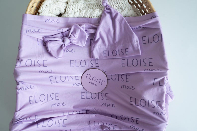 May include: A lavender baby blanket, headband, and hat set. The items are printed with the name "ELOISE" and "mae" in a repeating pattern. A round wooden sign with the same name is also included.