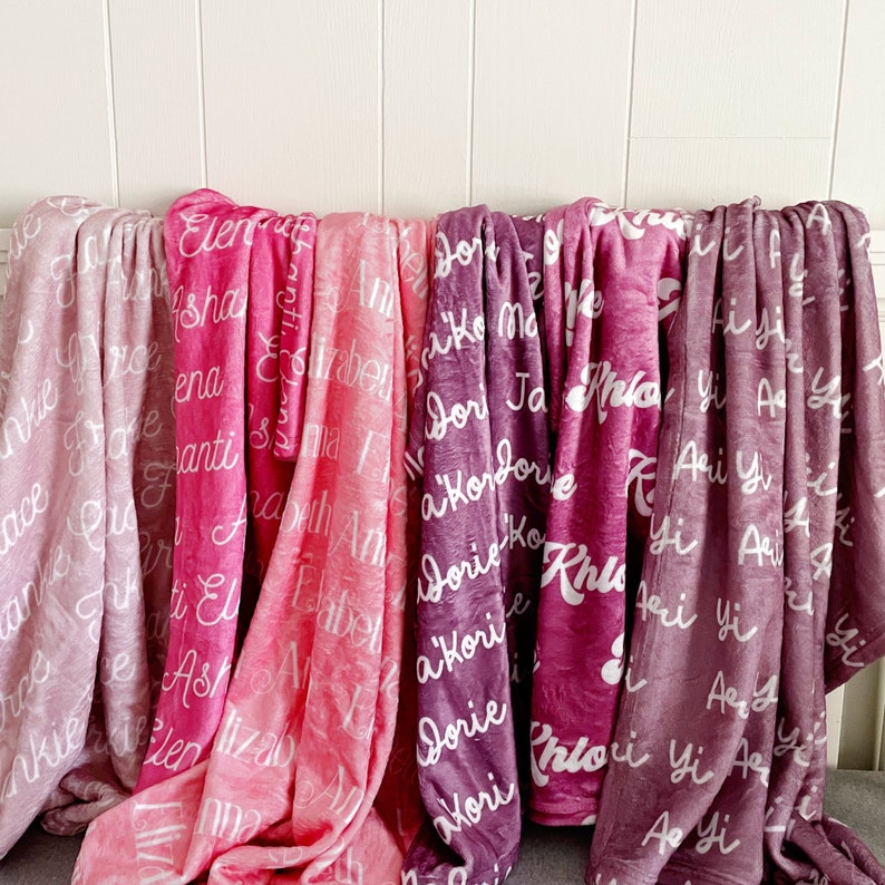 May include: A collection of soft, personalized blankets in various shades of pink and purple. Each blanket features a white script with names. The blankets are draped over a white surface, showcasing the different colors and designs.