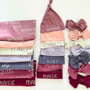 May include: A selection of baby accessories in shades of pink, peach, lavender, and burgundy. The image shows blankets, knotted hats, and headbands with bows. Each item is labelled with a name in white script.
