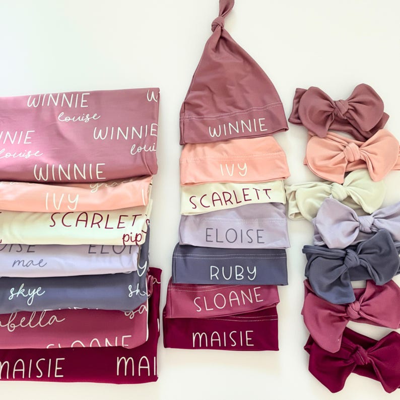 May include: A selection of baby accessories in shades of pink, peach, lavender, and burgundy. The image shows blankets, knotted hats, and headbands with bows. Each item is labelled with a name in white script.