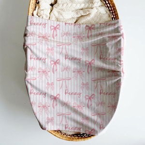 May include: A fabric cover featuring a repeating pattern of pink bows and vertical white stripes. The name Penny is printed in a cursive pink script throughout the design. The fabric is draped over a woven basket containing a white textured blanket.