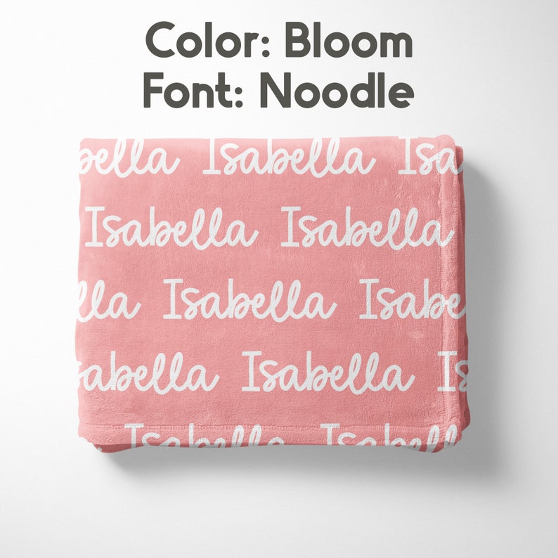 Peut inclure: Une couverture rose pli&eacute;e avec le nom "Isabella" r&eacute;p&eacute;t&eacute; en blanc dans une police cursive. La couleur de la couverture est "Bloom" et la police est "Noodle". La couverture est faite d'une mati&egrave;re douce.