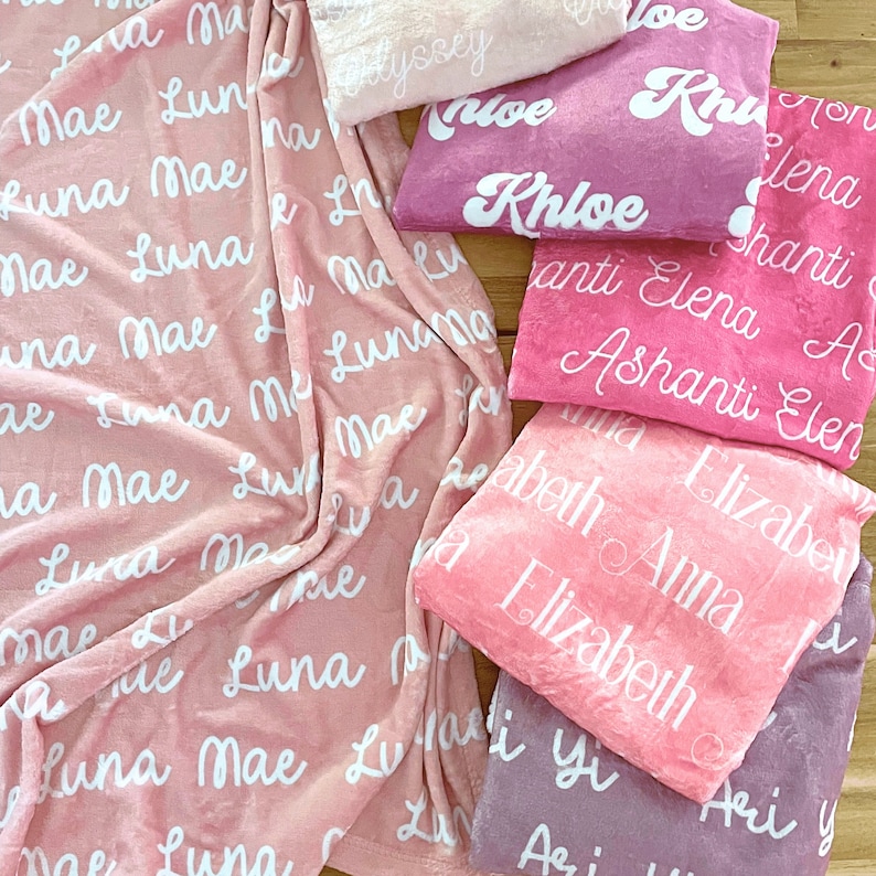 May include: A collection of personalized blankets in various shades of pink and purple. Each blanket features a name in a white, cursive font. The blankets are soft and plush, perfect for a baby or child.