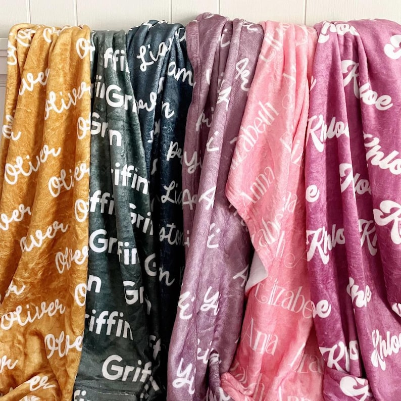 May include: A display of soft, personalized blankets in various colors, including gold, gray, purple, pink, and magenta. Each blanket features a different name in a white, cursive font, creating a cozy and custom feel.