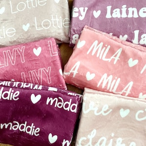 May include: A collection of personalized blankets in various shades of pink and purple. Each blanket features a name in white script font, accompanied by a small heart. The blankets are folded and arranged together, showcasing the soft texture.