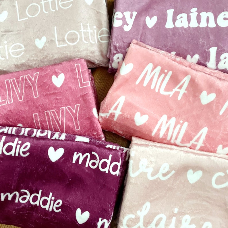 May include: A collection of personalized blankets in various shades of pink and purple. Each blanket features a name in white script font, accompanied by a small heart. The blankets are folded and arranged together, showcasing the soft texture.