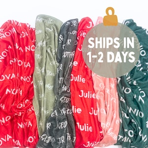 May include: A collection of soft blankets in various colors, including red, green, and gray. Each blanket is personalized with a name in white script. The blankets are folded and arranged together, creating a cozy display.