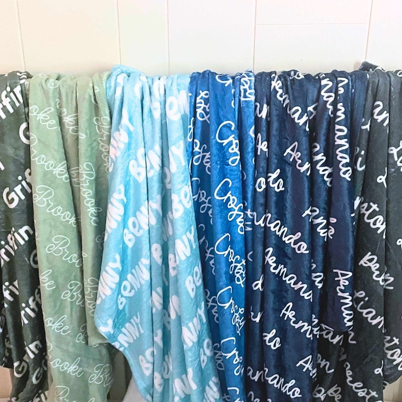 May include: A collection of soft, personalized blankets in various colors, including green, blue, and navy. Each blanket features a name in a white, cursive font, creating a custom and cozy feel. The blankets are displayed against a white background.