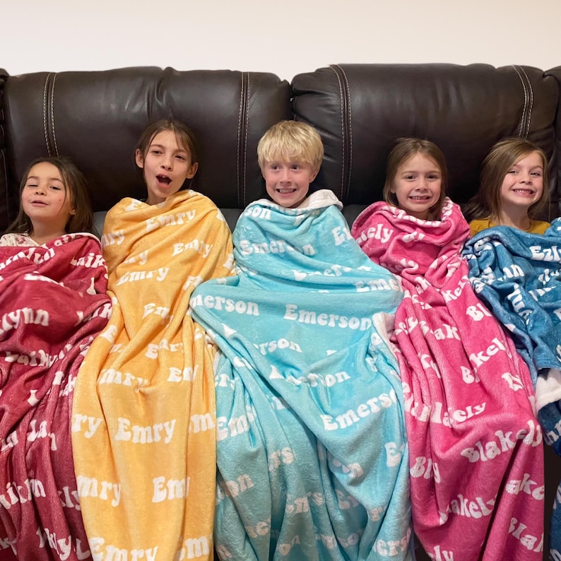 May include: Five children wrapped in personalized blankets of various colors, including red, yellow, blue, pink, and teal. Each blanket features a name printed in white, creating a cozy and personalized gift.