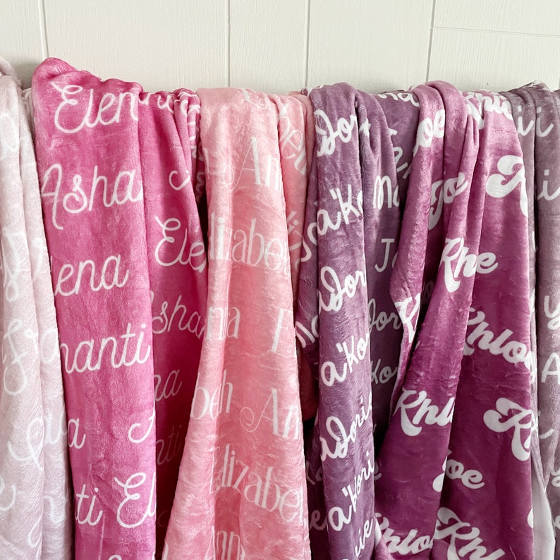 Peut inclure: Plusieurs couvertures douces et personnalis&eacute;es, dans des tons de rose et de violet. Chaque couverture affiche un nom diff&eacute;rent en blanc, en &eacute;criture cursive. Les couvertures sont drap&eacute;es sur une tringle blanche, sur fond de bois blanc.