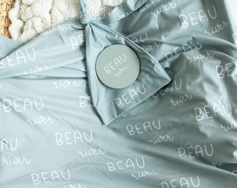 May include: A light blue baby blanket and matching knotted hat with the name "BEAU river" printed in white. A round wooden sign with the same name is also included. The blanket and hat are made of soft fabric.