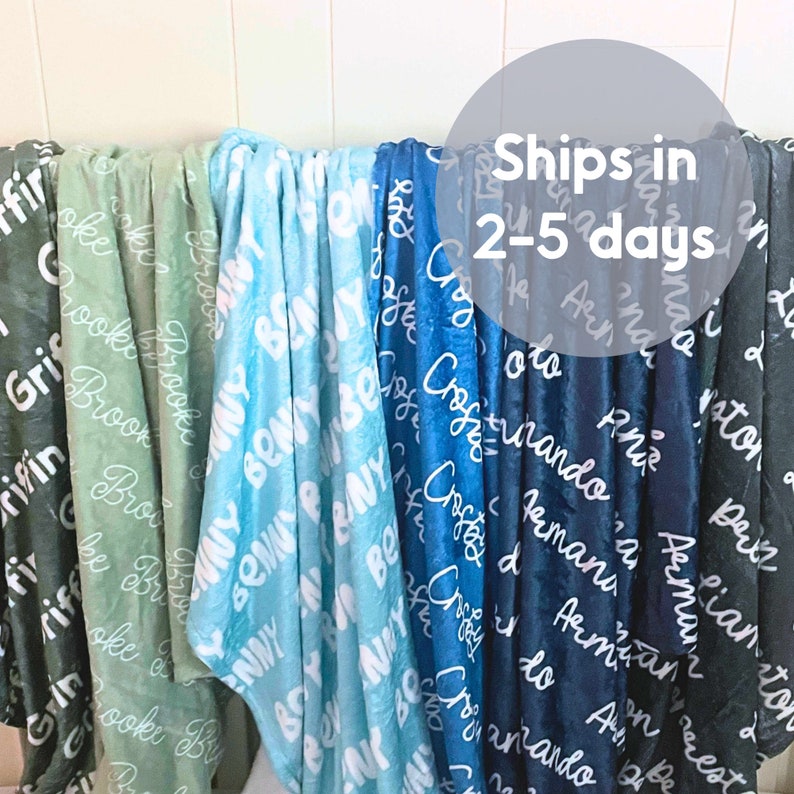 May include: A display of soft, personalized blankets in various colors, including green, blue, and navy. Each blanket is adorned with a name in a white, cursive font, creating a cozy and custom feel. The blankets are draped over a rod.