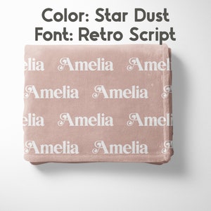 May include: A soft, star dust-colored blanket with the name "Amelia" repeated in white, using a retro script font. The blanket is folded neatly, showcasing the design. The text "Color: Star Dust" and "Font: Retro Script" are visible.