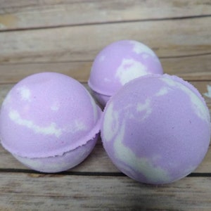 Puede incluir: Tres bombas de baño esféricas con un diseño veteado en lavanda y blanco. Las bombas de baño están dispuestas sobre una superficie de madera marrón claro. Tienen una forma suave y redondeada y una paleta de colores pastel.