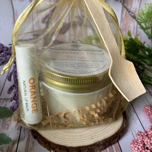 May include: A gold organza bag filled with brown shredded paper, a small white jar with a gold lid labeled "Gardener's Hand Scrub", and a white tube of lip balm labeled "ORANGE NATURAL LIP BALM".