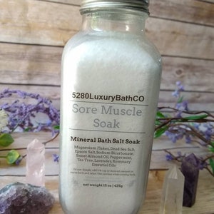 May include: A clear glass jar filled with white bath salts. The label reads "5280LuxuryBathCO Sore Muscle Soak Mineral Bath Salt Soak Magnesium Flakes, Dead Sea Salt, Epsom Salt, Sodium Bicarbonate, Sweet Almond Oil, Peppermint, Tea Tree, Lavender, Rosemary Essential Oils To use: Simply add 1/4 cup or desired amount to warm bath and relax. Use caution when exiting bath net weight 15 oz / 425g"